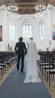 Wedding ceremony just for bride and groom in chapel. Action. Rear view of a man in suit and a woman in white dress walking together along empty rows in church towards the altar. video