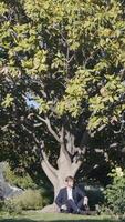 A man in a suit sitting under a huge tree. Action.A young man in a dark blue suit is sitting on the green grass under a huge tree with green leaves. video