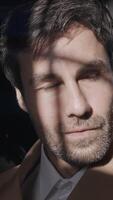 A handsome man with the sun on his face. Action.A bright illuminated portrait of a handsome man in a brown jacket sitting in a studio where people pose from behind. video
