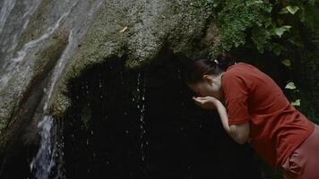 A beautiful mountain in which there is a small waterfall. Creative. A girl standing washing her face and drinking clean water in a cave through which a stream flows from a stream that runs in the cave video
