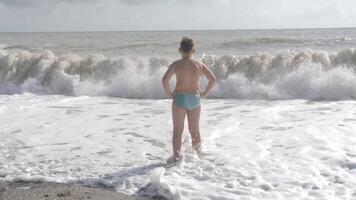 A boy who stands on the waves and admires the view of the sea. Creative . A boy in swimming shorts stands in the sea and looks at the sea waves. video