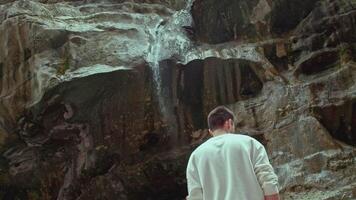 Rear view of man standing by mountain with flowing down small waterfall. Stock footage. Hiker enjoying beautiful nature. video