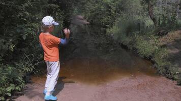 The child walks through the forest. CREATIVE. A boy in an orange T-shirt and brown pants runs in the forest. The guy walks through the forest next to the creek rear view video