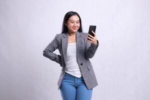 beautiful young office woman smile to mobile phone hold waist, holding cellphone wearing gray suit isolated on white background. for technology, lifestyle, shopping, transaction content photo