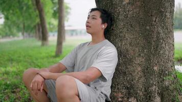 Asian man leaning on a tree thinking outside sitting on floor, He thinking about life, looking forward with hope and wearing AirPods while relaxing in a park, Healthy lifestyle, Relaxation. video