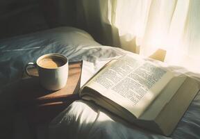 A cup of coffee on a bedside table next to an open book, with soft morning light illuminating the scene photo