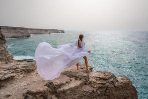 Happy freedom woman on the beach enjoying and posing in white dress over the sea. View of a girl in a fluttering white dress in the wind. Holidays, holidays at sea. photo