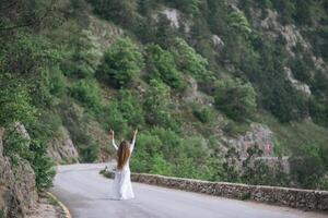 A woman is standing on a road, looking up at the sky. She is wearing a white dress and she is happy. The scene is set in a mountainous area, with trees and rocks in the background. photo