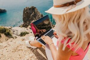 A woman is sitting on a rock by the ocean, typing on her laptop. Concept of relaxation and leisure, as the woman enjoys the beautiful view of the water while working on her computer. photo