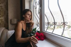 A woman in a black dress is sitting in front of a window with a bouquet of red roses. photo