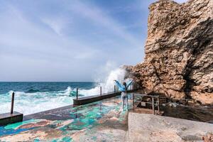 A woman in a blue jacket stands on a rock above a cliff above the sea and looks at the raging ocean. Girl traveler rests, thinks, dreams, enjoys nature. Peace and calm landscape, windy weather. photo