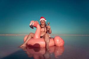 Santa Flamingo Christmas woman Riding Inflatable Flamingo Pink Lake. A woman wearing a Santa hat sits on a pink flamingo float in a body of water. He is smiling. photo