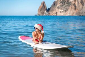 A woman in a red bikini and a Santa hat lies on a SUP board with the sea and a yacht floating on the horizon. He looks away and smiles. photo
