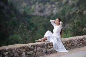 A woman in a white dress is sitting on a rock by a road. She is looking up at the sky. photo