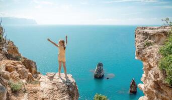 Happy girl stands on a rock high above the sea, wearing a yellow jumpsuit and sporting braided hair, depicting the idea of a summer vacation by the sea. photo