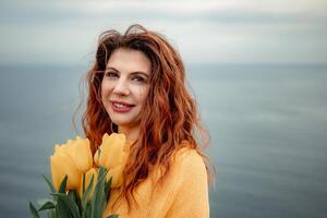 Portrait of a happy woman with hair flying in the wind against the backdrop of mountains and sea. Holding a bouquet of yellow tulips in her hands, wearing a yellow sweater photo