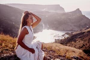 A woman in a white dress is sitting on a rock overlooking a body of water. She is enjoying the view and taking in the scenery. photo