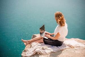 Freelance woman working on a laptop by the sea, typing away on the keyboard while enjoying the beautiful view, highlighting the idea of remote work. photo