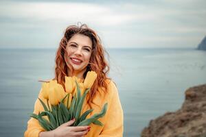 Portrait of a happy woman with hair flying in the wind against the backdrop of mountains and sea. Holding a bouquet of yellow tulips in her hands, wearing a yellow sweater photo