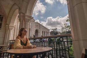 Woman Balcony View Architecture Tourist Attraction. A woman sits at a table on a balcony, looking out at a building and a cloudy sky. photo