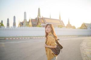 Traveler asian woman in her 30s, backpack slung over her shoulder, explores the intricate details of Wat Pra Kaew with childlike wonder. Sunlight dances on the golden rooftops. photo