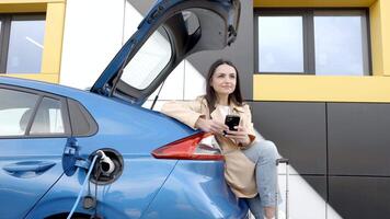 Jeune femme attendre mise en charge voiture batterie de petit Publique station et en utilisant téléphone intelligent tandis que mise en charge voiture video