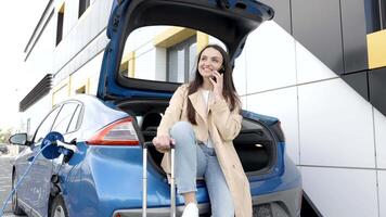 Jeune femme attendre mise en charge voiture batterie de petit Publique station et en utilisant téléphone intelligent tandis que mise en charge voiture video