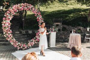 A woman stands in front of a large pink arch, singing to a crowd photo