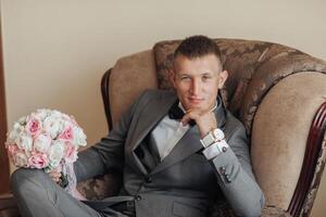 A man in a suit and tie sits in a chair with a bouquet of flowers in his lap photo