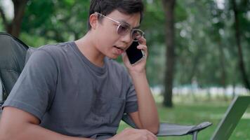 Programmer sit on a chair in a park use laptop discuss project details with colleague by phone, call having formal conversation provide help to client, distance communication, modern tech concept. video