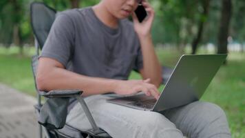 Businessman use smartphone and laptop sit on a chair in a park, Programmer hand hold smartphone conversation with abroad client distance meeting and typing on a laptop, Working online. video