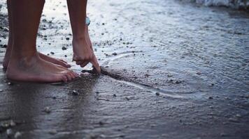 Hands delicately trace a heart on the soft sand near the water, creating a simple yet meaningful expression. The heart, shaped with care, captures the essence of love as it rests by the shore. video