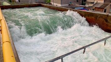 Rapid Water Flow in a Concrete Channel. A view of a water discharge channel, with water rapidly flowing through it. The concrete channel is surrounded by industrial buildings and pipes. video