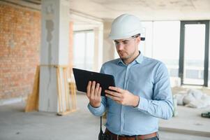 Supervisor standing in building in construction process, holding tablet and checking on works photo