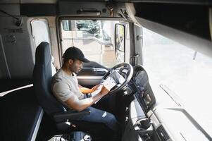 African-American truck driver is sitting behind the wheel holding a clipboard and checking the documents for the cargo photo