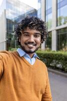 Smiling man taking selfie outside modern office building. Displays confidence and casual style with a sweater and shirt. Enjoys outdoor urban setting during a sunny day, capturing joyful photo