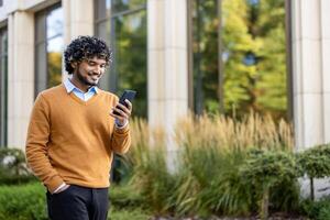 Confident man standing outside modern office building, holding phone. Wearing casual orange sweater, enjoying sunny day. Reflection of trees seen on glass windows, creating calm serene atmosphere. photo