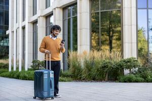 Smiling business traveler using smartphone while pulling suitcase in urban setting. Person wearing orange sweater walking outside office building. Captures concept of business travel photo