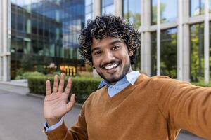 Cheerful man taking selfie outside modern office building. Man is smiling, waving, creating welcoming atmosphere. Bright sunny day enhances positive and professional mood. photo