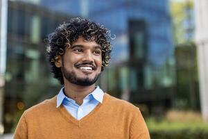Confident Indian businessman smiling in urban environment, wearing orange sweater and blue shirt. Depicts professional success and positive outlook. Concepts of business confidence, optimism photo