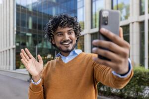 Cheerful man taking selfie with phone in city environment, wearing casual clothes. Captures joyful expression, waving to camera, experiencing happiness and connection. Ideal for themes of technology, communication, and modern lifestyle. photo