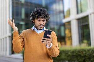 Man with curly hair standing outside modern office building looks at phone with frustration. Wearing a brown sweater, he expresses confusion or dissatisfaction with technology issues. photo