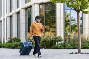 Man dressed in casual clothing walking with suitcase, using smartphone outside modern office building. Represents travel, technology, and connectivity in urban environment. photo