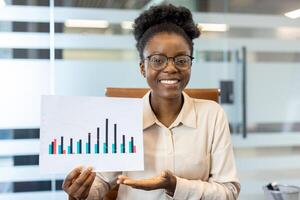 Young African American businesswoman at workplace holding data chart. Confident employee presenting statistical graph in modern office setting while smiling at camera. photo