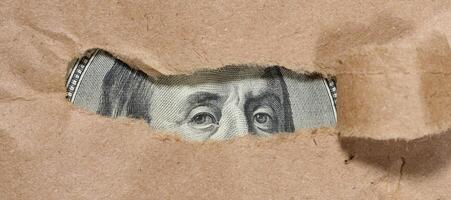 A piece of a hundred dollar bill is visible through a hole in the paper. Symbolism of hidden or concealed wealth, money that remains out of sight, or financial resources. photo