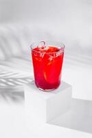 Refreshing red cocktail standing on white cube with tropical palm leaf shadow photo