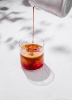 Espresso pouring into glass with red liquid and ice cubes on white background with shadows photo