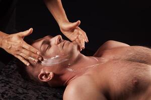 A man is getting a facial treatment at a spa. He is lying on a dark surface with his eyes closed, and the masseur is applying cream to his face with gentle hands. Facial massage concept photo