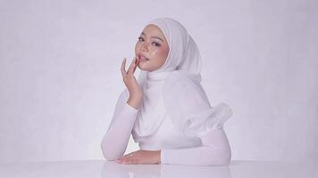 portrait sidewise Beautiful young hijab girl asia pouting to camera over hands resting on white table and touching cheeks isolated on white background video