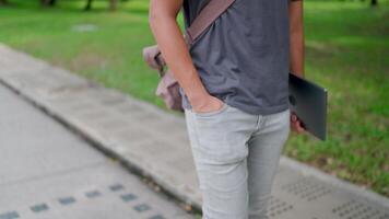 Man is walking with his hands in his pants and holding a laptop in the park, working online, He is walking in nature to relax from work and study, studying online, modern tech concept. video
